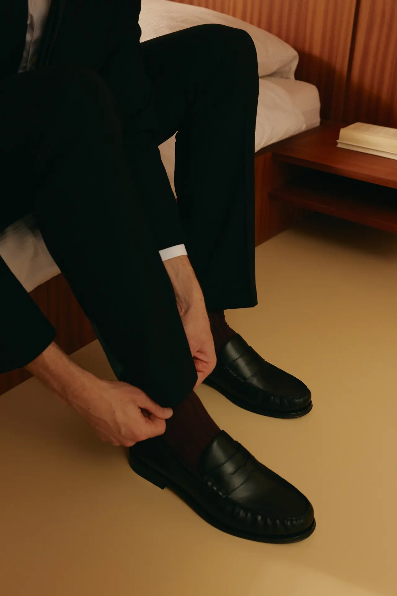 Person in a black suit adjusting a trouser cuff while seated beside a bed.